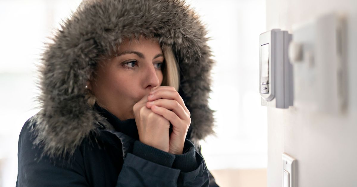 Woman wearing a large winter coat, blowing on her hands and looking at her furnace's thermostat.